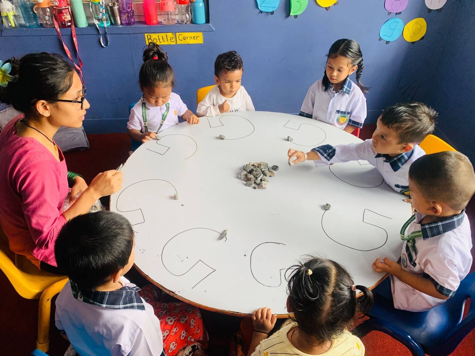 A teacher and eight toddlers around a low round table, tracing letter shapes with pebbles