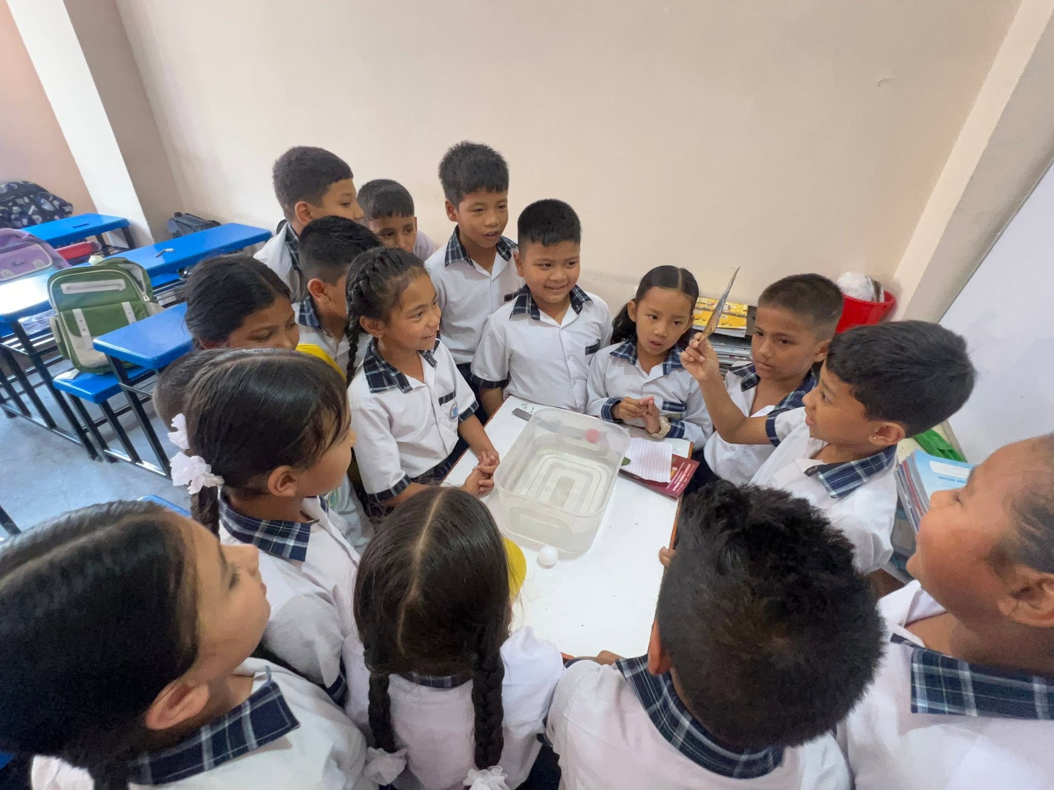 Primary pupils in checked-collar uniforms crowded around a classroom table watching a water experiment